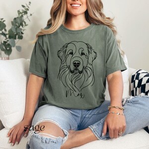 Op de afbeelding: Een vrouw draagt een saliegroen T-shirt met een zwarte lijntekening van een hondenkop. De naam van de hond, "Max", staat onder de tekening. De vrouw draagt een blauwe jeans en zit op een witte bank.