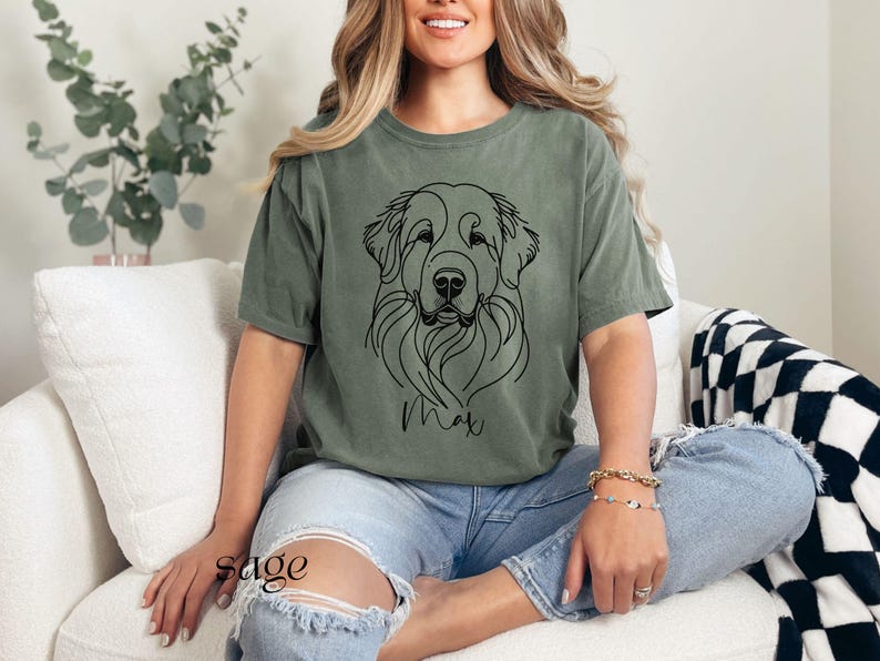 Op de afbeelding: Een vrouw draagt een saliegroen T-shirt met een zwarte lijntekening van een hondenkop. De naam van de hond, "Max", staat onder de tekening. De vrouw draagt een blauwe jeans en zit op een witte bank.