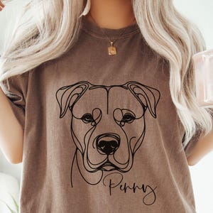 May include: A brown t-shirt featuring a black line drawing of a dog's face with the name "Penny" in script. The person is holding a pink mug with the words "cool mom club" in white. A teal scrunchie is on the person's wrist.
