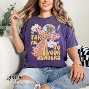Puede incluir: Camiseta morada con un dise&ntilde;o retro de un extraterrestre con sombrero de vaquero dentro de un OVNI, con el texto "Take Me To Your Readers". El dise&ntilde;o incluye nubes, estrellas y un rayo arco&iacute;ris. La camiseta se usa con vaqueros azules.