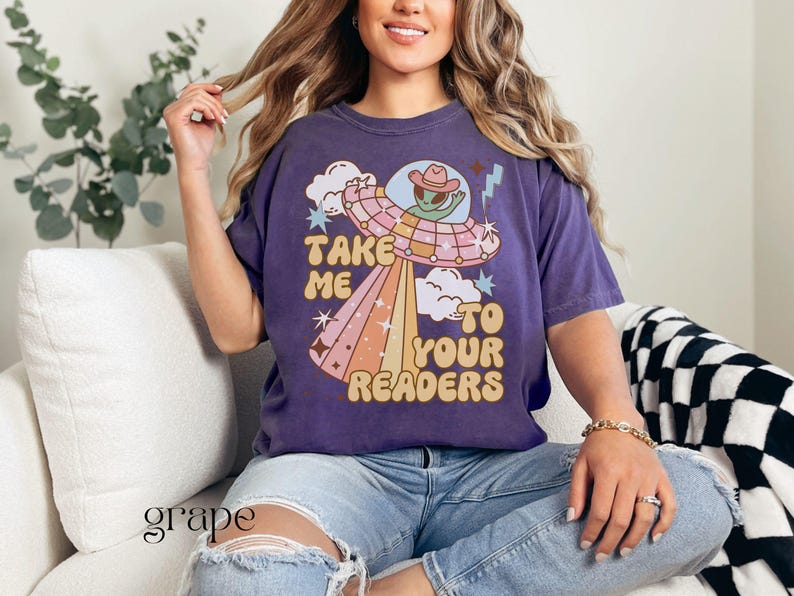 Puede incluir: Camiseta morada con un dise&ntilde;o retro de un extraterrestre con sombrero de vaquero dentro de un OVNI, con el texto "Take Me To Your Readers". El dise&ntilde;o incluye nubes, estrellas y un rayo arco&iacute;ris. La camiseta se usa con vaqueros azules.