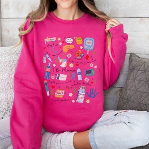 Op de afbeelding: Een roze sweatshirt met een grafisch ontwerp met verschillende diabetesgerelateerde items, waaronder een bloedglucosemeter, een insulinepomp en spuiten. De tekst "T1D Mama" staat op het sweatshirt.