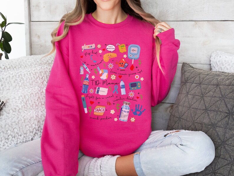 Op de afbeelding: Een roze sweatshirt met een grafisch ontwerp met verschillende diabetesgerelateerde items, waaronder een bloedglucosemeter, een insulinepomp en spuiten. De tekst "T1D Mama" staat op het sweatshirt.