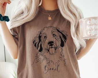 May include: A brown t-shirt featuring a black line drawing of a dog's face with the name "Basil" in script. The person is also wearing a green scrunchie and holding a pink mug with white text.