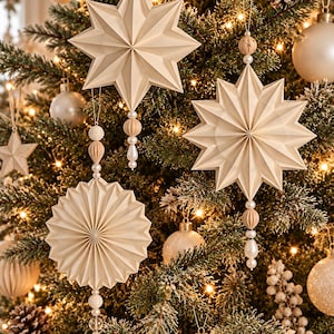May include: Three cream-colored paper ornaments hanging from a Christmas tree. Two are multi-pointed stars and one is a circular accordion-folded fan. Each ornament features a vertical string of decorative wooden and pearl-like beads.