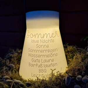 May include: A glowing white lantern with the word "Sommer" and other German words etched on its surface. The lantern is surrounded by moss and small plants. The lantern has a handle at the top.