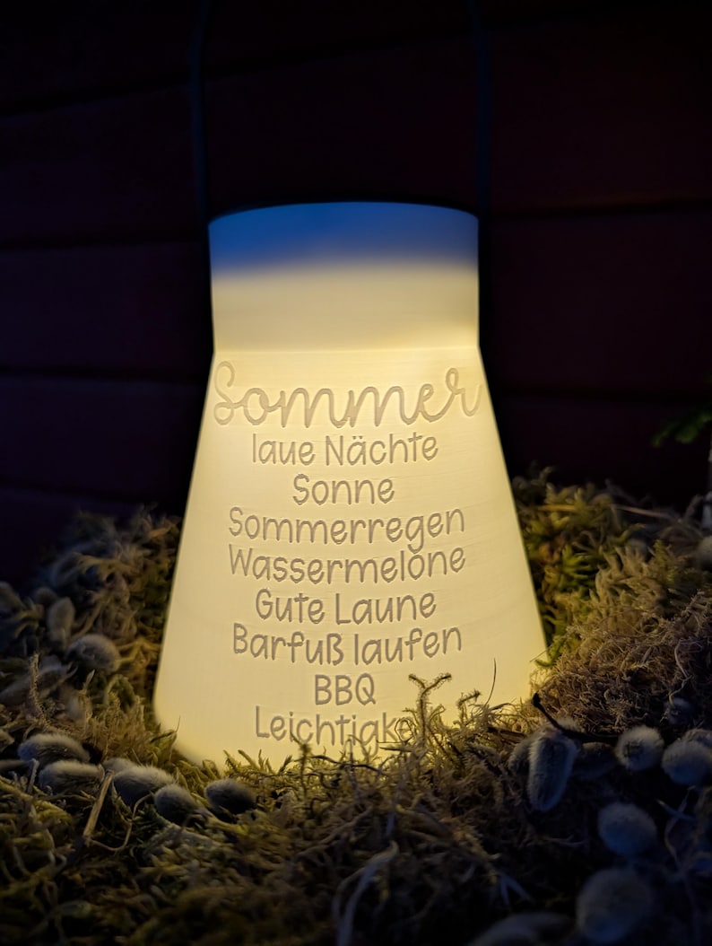 May include: A glowing white lantern with the word "Sommer" and other German words etched on its surface. The lantern is surrounded by moss and small plants. The lantern has a handle at the top.