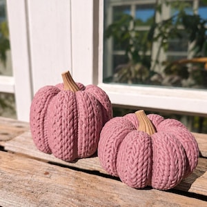 May include: Two decorative pumpkins in a dusty rose pink color, featuring a textured knit pattern across the surface and a light brown, ribbed stem at the top. The pumpkins are rounded with deep vertical grooves, mimicking the appearance of yarn.