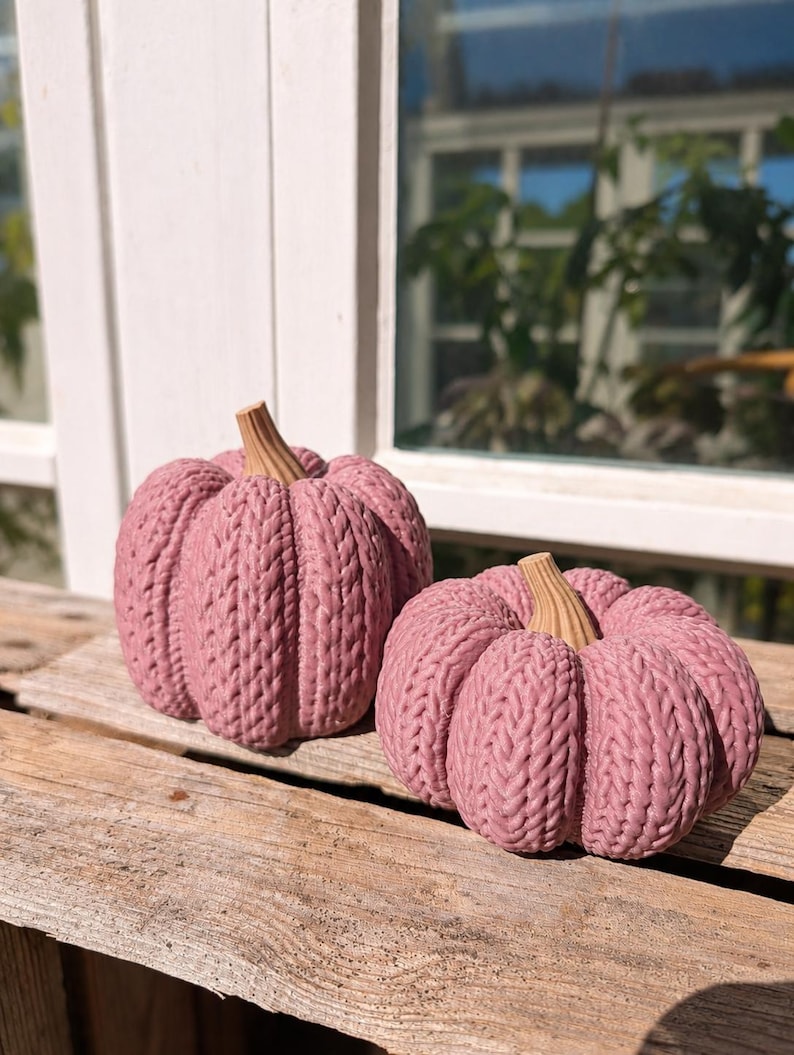 May include: Two decorative pumpkins in a dusty rose pink color, featuring a textured knit pattern across the surface and a light brown, ribbed stem at the top. The pumpkins are rounded with deep vertical grooves, mimicking the appearance of yarn.
