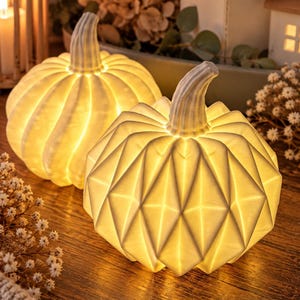 May include: Two off-white pumpkin-shaped lamps with internal warm lighting. One pumpkin features a smooth, ribbed surface, while the other displays a geometric, faceted texture. Both have a prominent stem and emit a soft, golden glow.