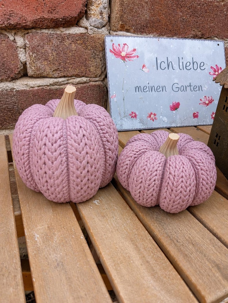 May include: Two dusty pink pumpkins with a textured knit pattern and light beige stems. Behind the pumpkins is a rectangular metal sign with a floral design and the German text "Ich liebe meinen Garten" printed in a dark gray font.