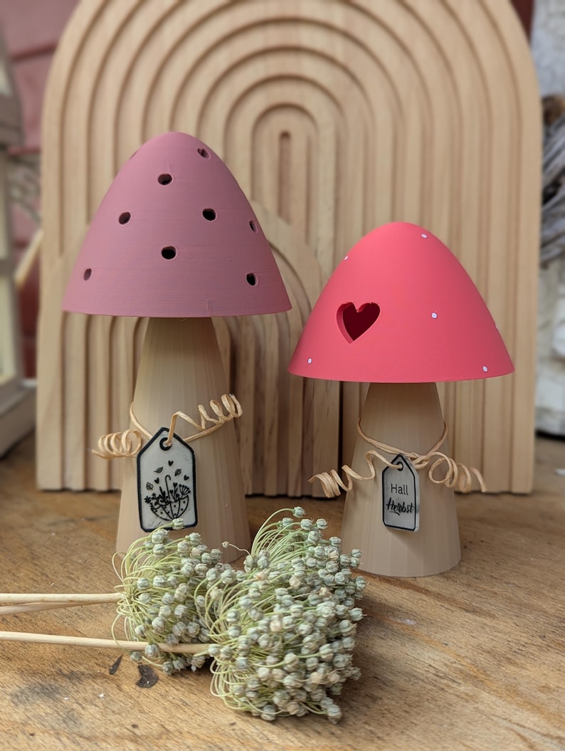 May include: Two wooden mushroom figurines with pink and coral caps. The pink mushroom has small holes, while the coral one has a heart-shaped cutout. Each has a tag with text and is tied with twine. A bundle of dried flowers rests in front.