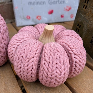 May include: A round, dusty pink pumpkin decoration featuring a textured, knit-like surface pattern and a light tan, ridged stem. The object is segmented into several distinct, plump sections, creating a soft, rounded appearance throughout.