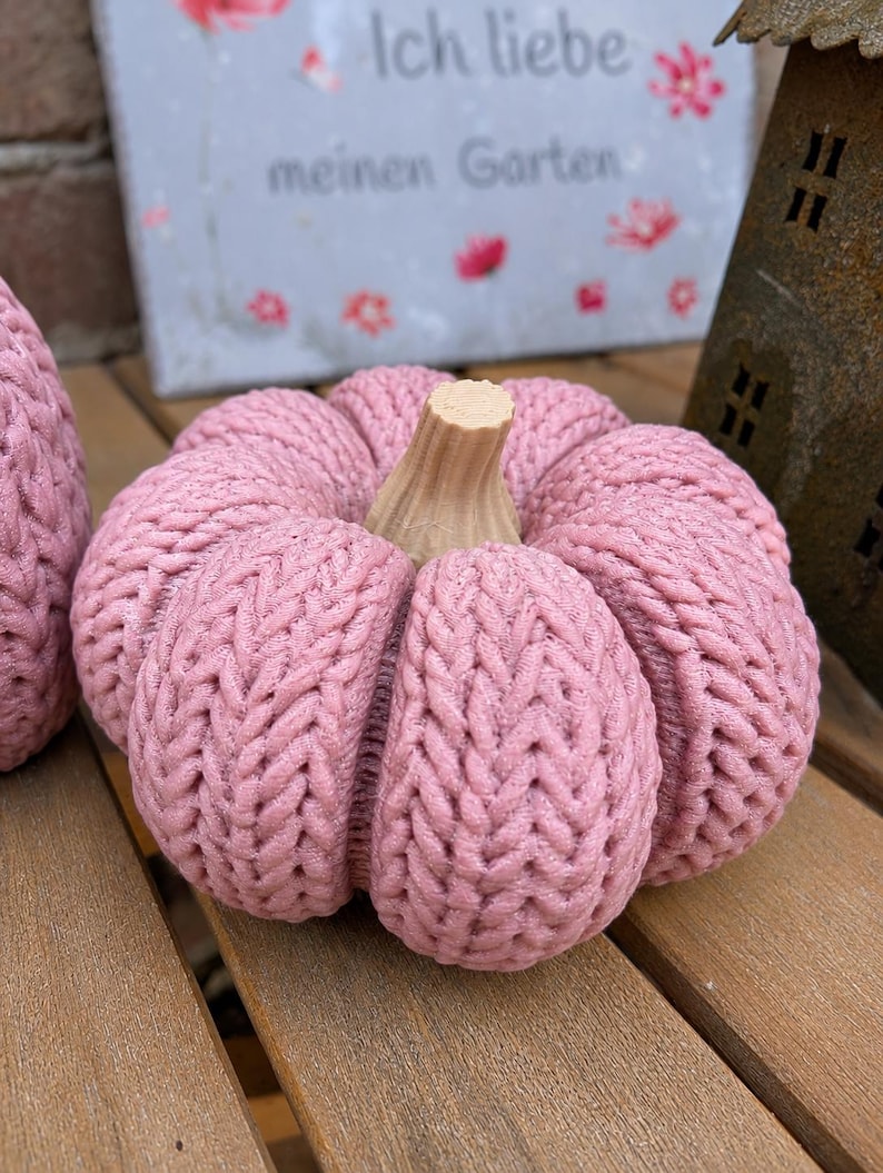 May include: A round, dusty pink pumpkin decoration featuring a textured, knit-like surface pattern and a light tan, ridged stem. The object is segmented into several distinct, plump sections, creating a soft, rounded appearance throughout.