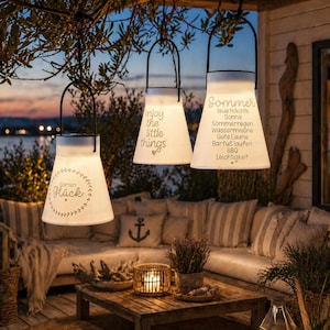 May include: Three hanging white lanterns with black handles, each with text. One says "enjoy the little things", another "Sommer", and the third "Garten gl&uuml;ck". The lanterns are hanging outside near a seating area.