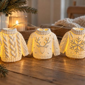 Puede incluir: Una guirnalda de tres luces LED blancas con forma de jerséis en miniatura. Cada jersey presenta un patrón único: uno con trenzas, otro con un motivo de reno y otro con un diseño de copo de nieve, todos emitiendo una luz cálida.