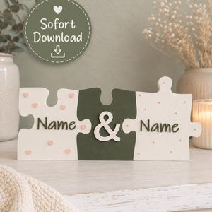 Puede incluir: Un letrero decorativo de madera con forma de piezas de rompecabezas que incluye dos piezas exteriores de color crema con pequeños corazones rosas y una pieza central verde oscuro. El texto "Name & Name" aparece en letras oscuras. Una insignia circular dice "Sofort Download".