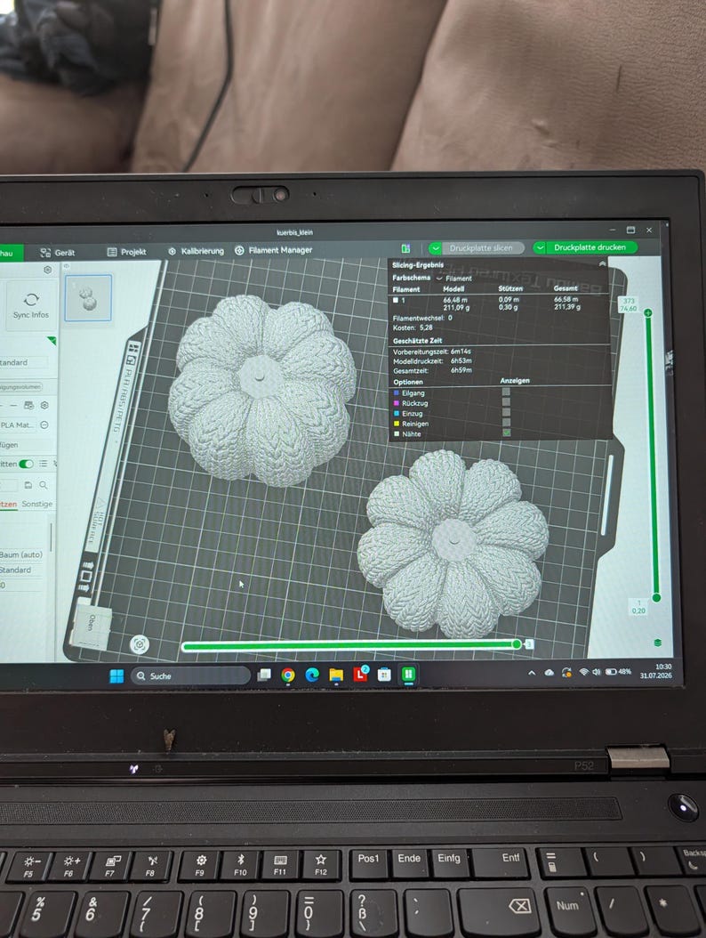 May include: A laptop screen displaying a 3D slicing software interface with two light gray pumpkin-shaped models on a digital build plate. The software window shows technical data including filament usage, print time, and various settings in German.