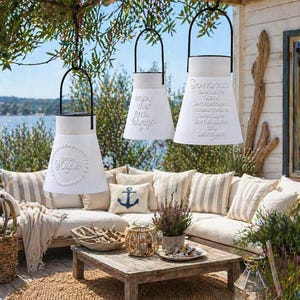 May include: Three white, cone-shaped lanterns with black handles hang outdoors. One lantern reads "Garden Gl&uuml;ck", another "enjoy the little things", and the third "Sommer". The lanterns are part of an outdoor patio setting with a sofa, coffee table, and rug.