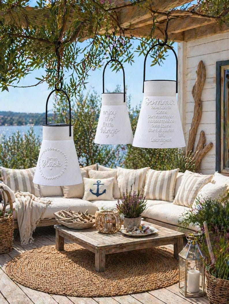 May include: Three white, cone-shaped lanterns with black handles hang outdoors. One lantern reads "Garden Gl&uuml;ck", another "enjoy the little things", and the third "Sommer". The lanterns are part of an outdoor patio setting with a sofa, coffee table, and rug.