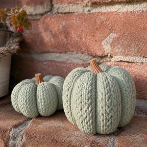 May include: Two sage green decorative pumpkins featuring a textured, knitted cable pattern and a brown stem. The pumpkins are rounded with deep vertical ridges, mimicking the appearance of a classic gourd, and are crafted with a uniform knit finish.