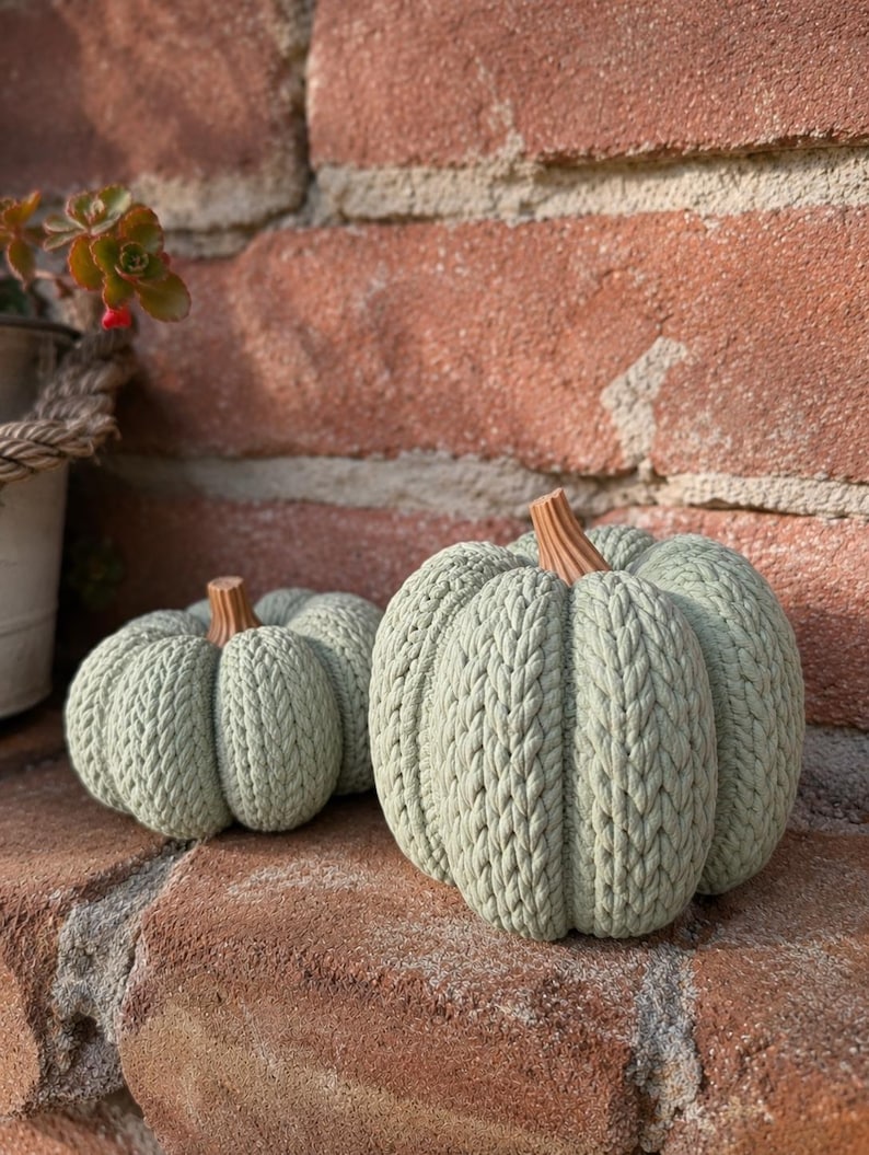 May include: Two sage green decorative pumpkins featuring a textured, knitted cable pattern and a brown stem. The pumpkins are rounded with deep vertical ridges, mimicking the appearance of a classic gourd, and are crafted with a uniform knit finish.