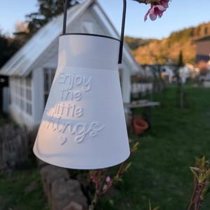 May include: White, bell-shaped decorative hanging planter with a black handle. The planter has the embossed text "Enjoy the little things" and two small hearts. The background includes a small white house and a flowering tree branch.