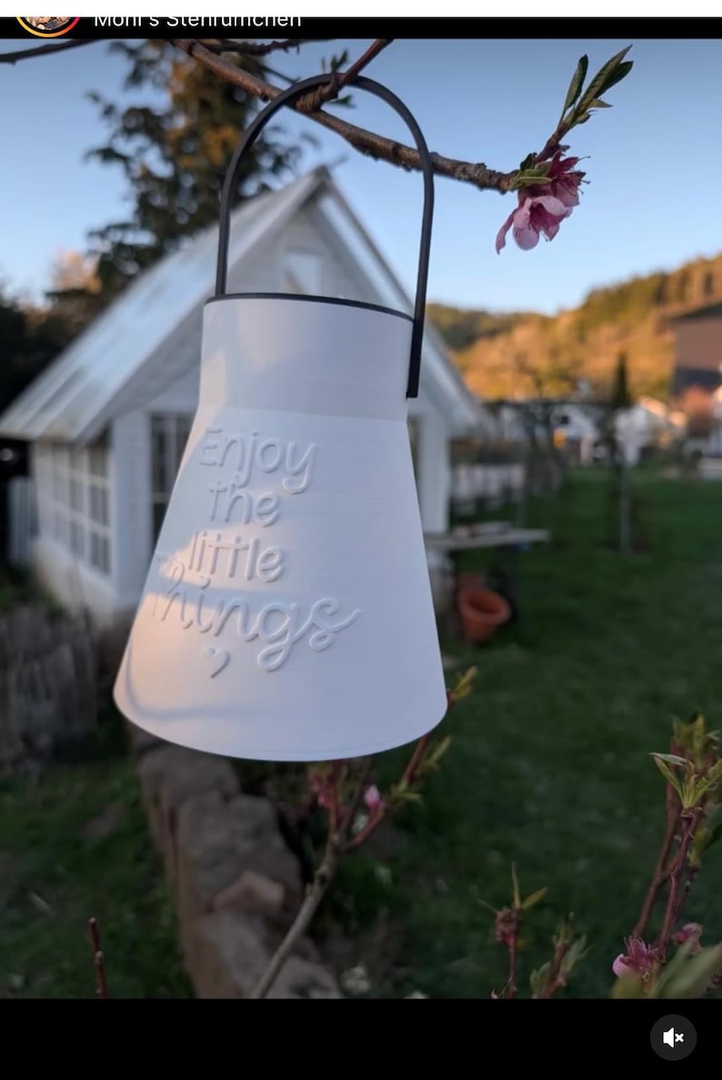 May include: White, bell-shaped decorative hanging planter with a black handle. The planter has the embossed text "Enjoy the little things" and two small hearts. The background includes a small white house and a flowering tree branch.