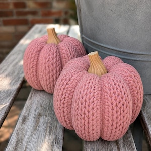 May include: Two round, dusty pink pumpkins crafted with a thick, textured knit yarn pattern. Each pumpkin features a smooth, light brown wooden stem with vertical ridges, creating a distinct contrast against the soft, ribbed fabric body.