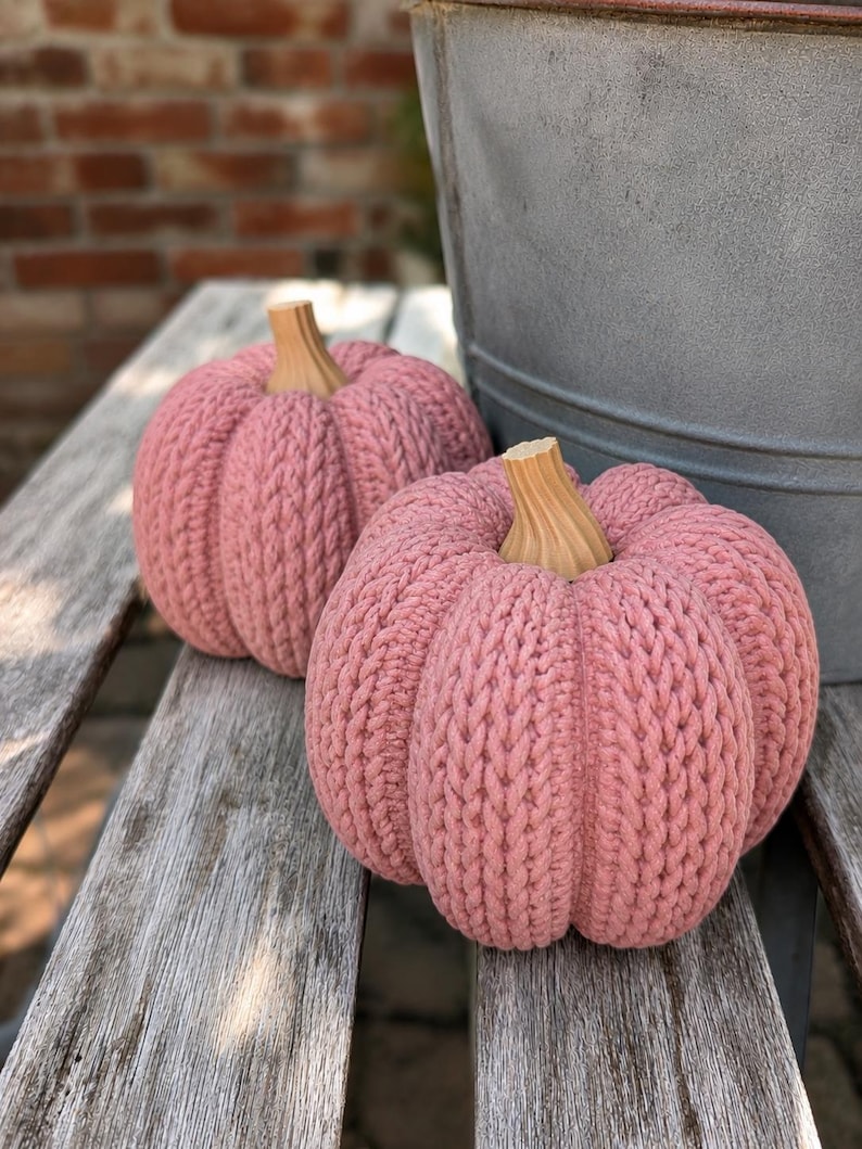 May include: Two round, dusty pink pumpkins crafted with a thick, textured knit yarn pattern. Each pumpkin features a smooth, light brown wooden stem with vertical ridges, creating a distinct contrast against the soft, ribbed fabric body.