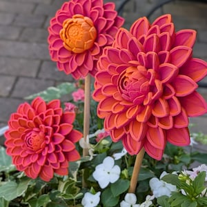 May include: Three decorative dahlia flower sculptures featuring layered petals in vibrant shades of pink and orange. Each flower is attached to a slender wooden stake, positioned among green foliage and small white flowers in a garden setting.