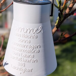 May include: A white, bell-shaped lantern with a black handle and rim, hanging from a tree branch. The lantern has embossed text in German, including "Sommer" and other summer-related words. The background is blurred with green and pink hues.
