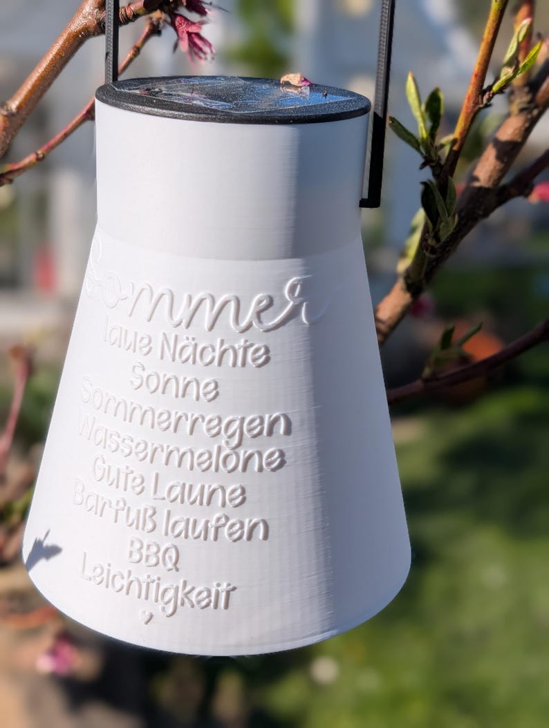 May include: A white, bell-shaped lantern with a black handle and rim, hanging from a tree branch. The lantern has embossed text in German, including "Sommer" and other summer-related words. The background is blurred with green and pink hues.
