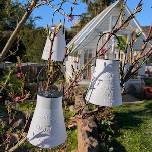 May include: Three white solar-powered lanterns with black handles hang from a tree branch. The lanterns have a milk jug shape and feature embossed text. A white greenhouse is visible in the background, with a blue sky.