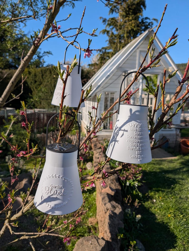 May include: Three white solar-powered lanterns with black handles hang from a tree branch. The lanterns have a milk jug shape and feature embossed text. A white greenhouse is visible in the background, with a blue sky.