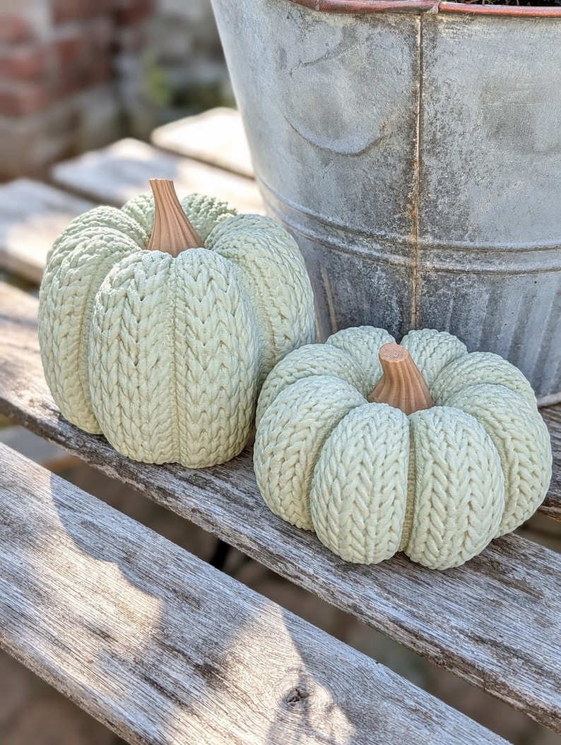 May include: Two decorative pumpkins featuring a light sage green color and a textured surface that mimics a knitted cable stitch pattern. Each pumpkin is topped with a smooth, light brown stem that has vertical ridges extending from the center.