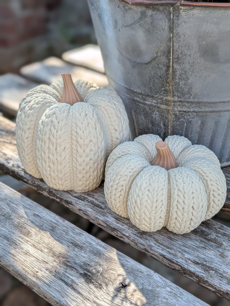 May include: Two off-white decorative pumpkins featuring a textured cable knit pattern across their rounded surfaces. Each pumpkin is topped with a smooth, light brown stem. The pumpkins are positioned side by side, displaying a detailed ribbed design.