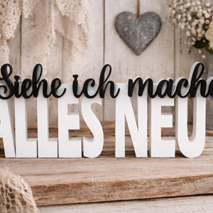 Könnte beinhalten: Ein dekoratives Holzschild mit dem deutschen Satz "Siehe ich mache ALLES NEU" in Schwarz und Weiß. Das Schild steht auf einer Holzoberfläche, mit einer brennenden Kerze, Blumen und einer herzförmigen Dekoration im Hintergrund.