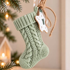 Pode incluir: Um enfeite de meia de Natal em tricô verde sálvia com padrão de tranças e punho canelado. Uma etiqueta branca em forma de estrela com o número 1 impresso em verde escuro está presa à meia com uma fita bege e um cordão de juta.