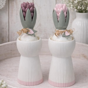 Könnte beinhalten: Zwei dekorative Blumengestecke. Jedes hat eine weiße, geriffelte Vase mit rosa Basis, gekrönt von einer Kunstblume. Eine Blume ist lila, die andere rosa, beide mit grünen Akzenten und Bindfaden.