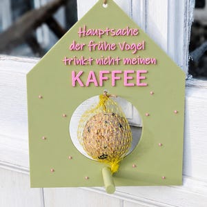 Könnte beinhalten: Ein grünes Vogelhaus in Hausform mit dem deutschen Text "Hauptsache der frühe Vogel trinkt nicht meinen KAFFEE" in Rosa. Durch eine kreisförmige Öffnung ist ein runder Vogelfutterball zu sehen. Das Vogelhaus wird von einer Schnur gehalten.