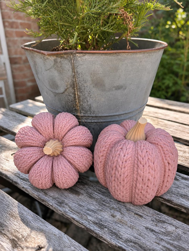 May include: Two decorative pumpkins in a dusty pink shade, featuring a textured knit pattern across the surface and a light-colored, ribbed stem at the top. The pumpkins are rounded and segmented, displaying a soft, matte finish throughout.