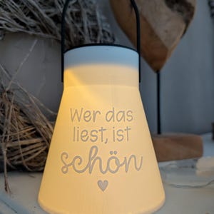 May include: A white ceramic lantern with a black handle. The lantern has the words "Wer das liest, ist sch&ouml;n" in a script font, with a small heart symbol. The lantern is lit from within, creating a warm glow.