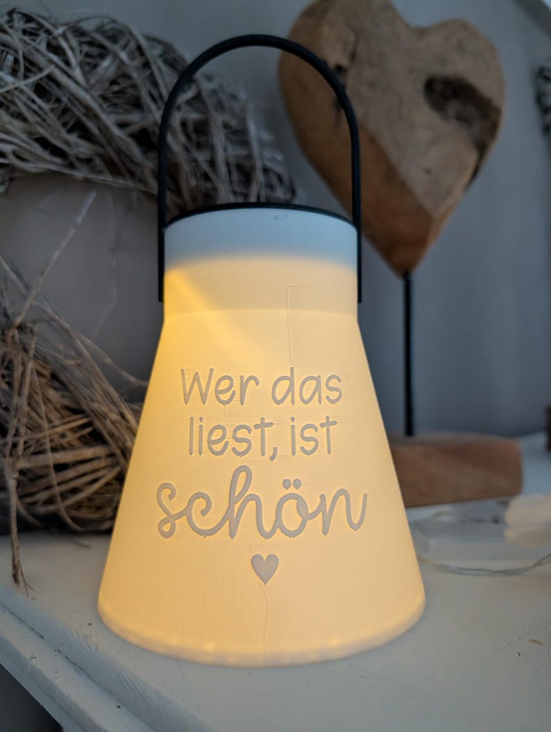 May include: A white ceramic lantern with a black handle. The lantern has the words "Wer das liest, ist sch&ouml;n" in a script font, with a small heart symbol. The lantern is lit from within, creating a warm glow.