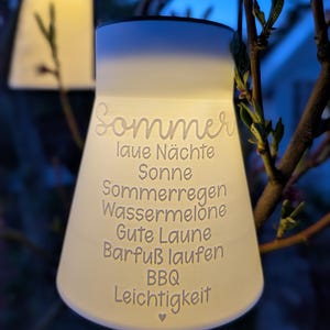 May include: A glowing, white, bell-shaped outdoor lantern with the word "Sommer" and other German words etched on its surface. The lantern is hanging from a tree branch, with a soft, warm light emanating from within. The background is a blurred view of a dark blue sky.