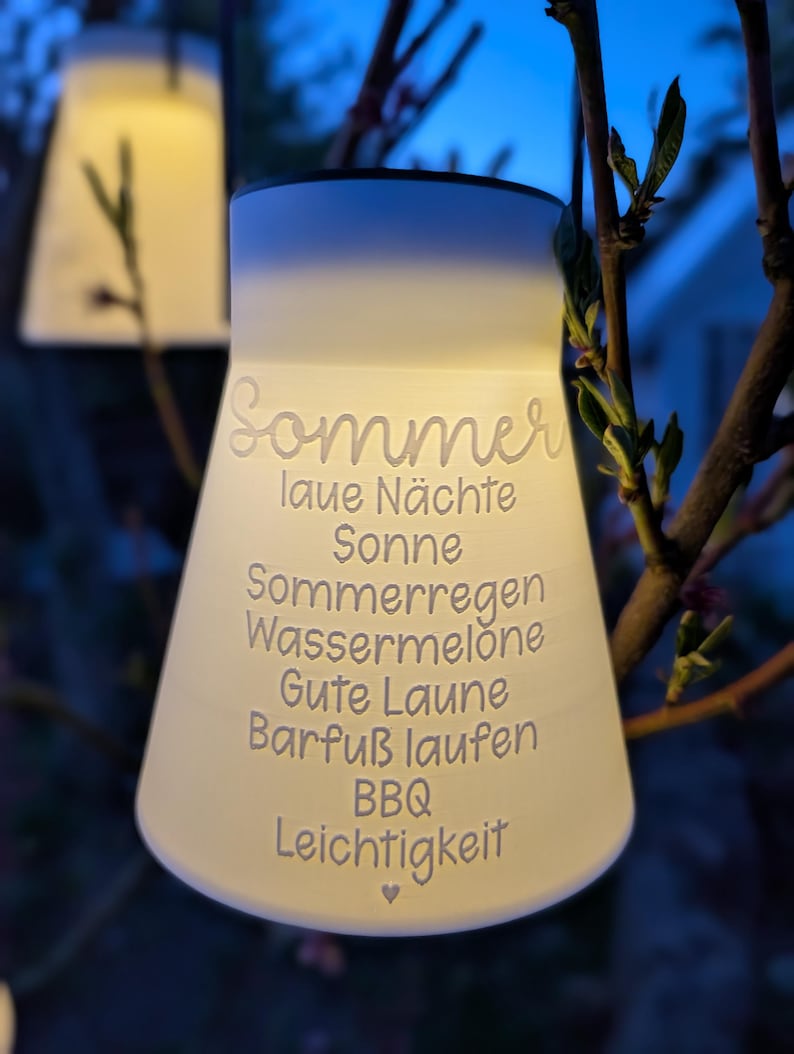 May include: A glowing, white, bell-shaped outdoor lantern with the word "Sommer" and other German words etched on its surface. The lantern is hanging from a tree branch, with a soft, warm light emanating from within. The background is a blurred view of a dark blue sky.