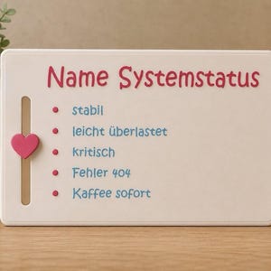 May include: A white rectangular sign with the words "Name Systemstatus" in red. A pink heart-shaped slider is on the left. Below the title are bullet points with German words: "stabil", "leicht überlastet", "kritisch", "Fehler 404", and "Kaffee sofort".