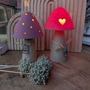 May include: Two decorative mushroom-shaped lamps with glowing caps. One has a purple cap with yellow dots, the other a red cap with a heart-shaped cutout. Both have beige stems and are on a wooden surface. The text "Hall Harvest" is on a tag.