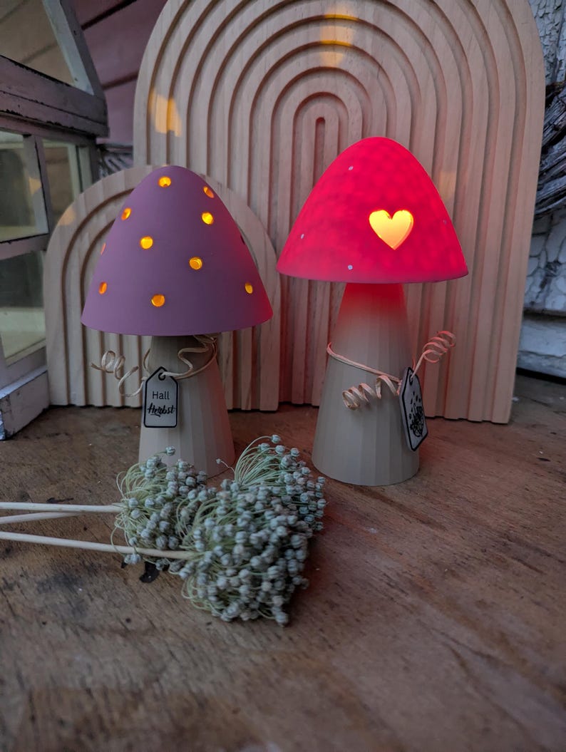 May include: Two decorative mushroom-shaped lamps with glowing caps. One has a purple cap with yellow dots, the other a red cap with a heart-shaped cutout. Both have beige stems and are on a wooden surface. The text "Hall Harvest" is on a tag.