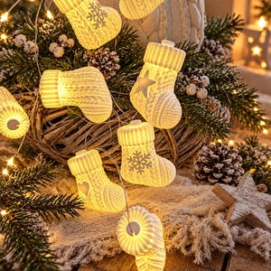 Puede incluir: Una cadena de luces LED con pequeñas fundas blancas en forma de calcetín con textura de punto. Cada calcetín está iluminado desde el interior y decorado con varias formas en relieve, incluyendo copos de nieve, estrellas y corazones, unidos por un cable.